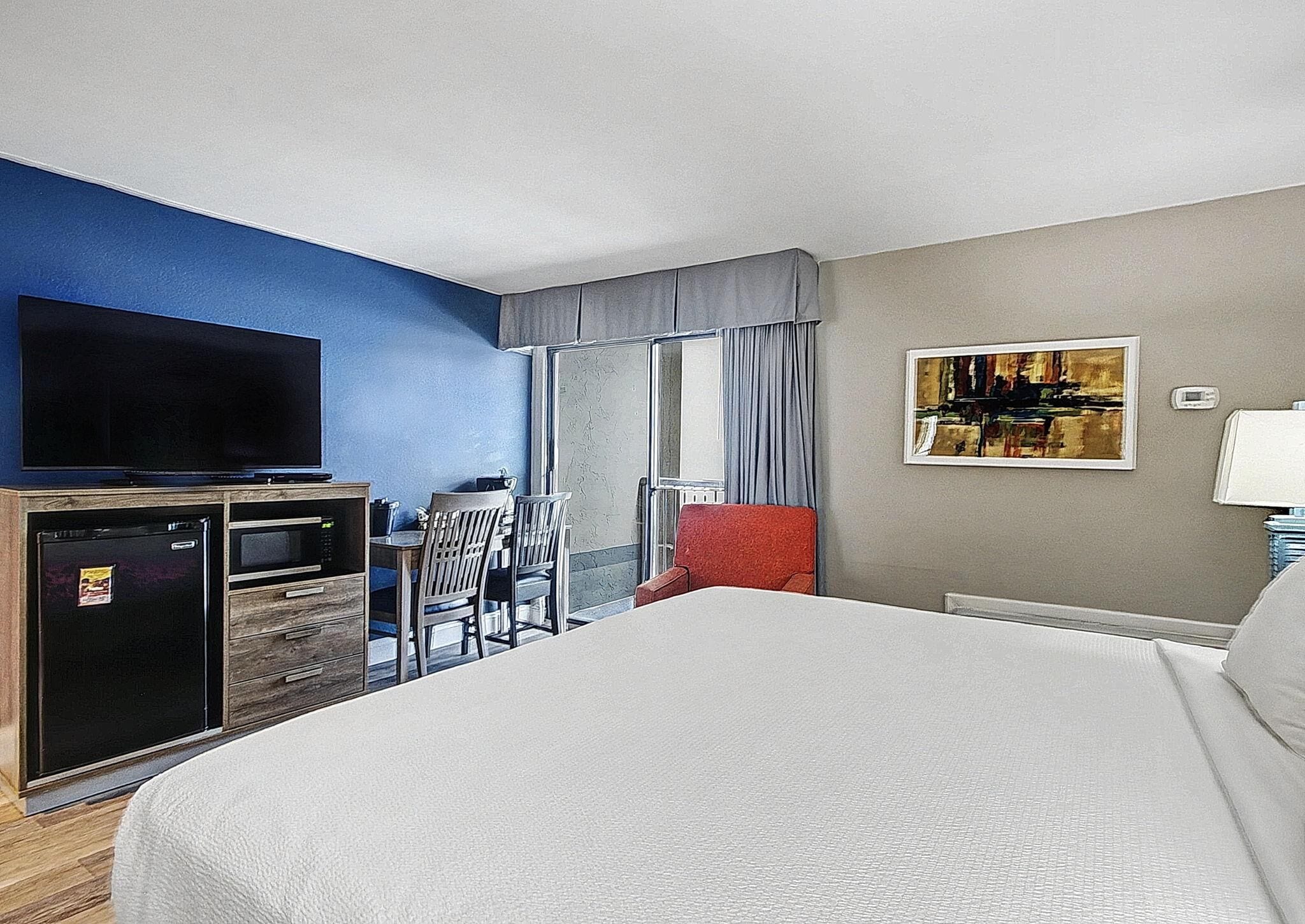 A hotel room with a bed, a TV, a small kitchenette, a window with curtains, and a painting on the wall.