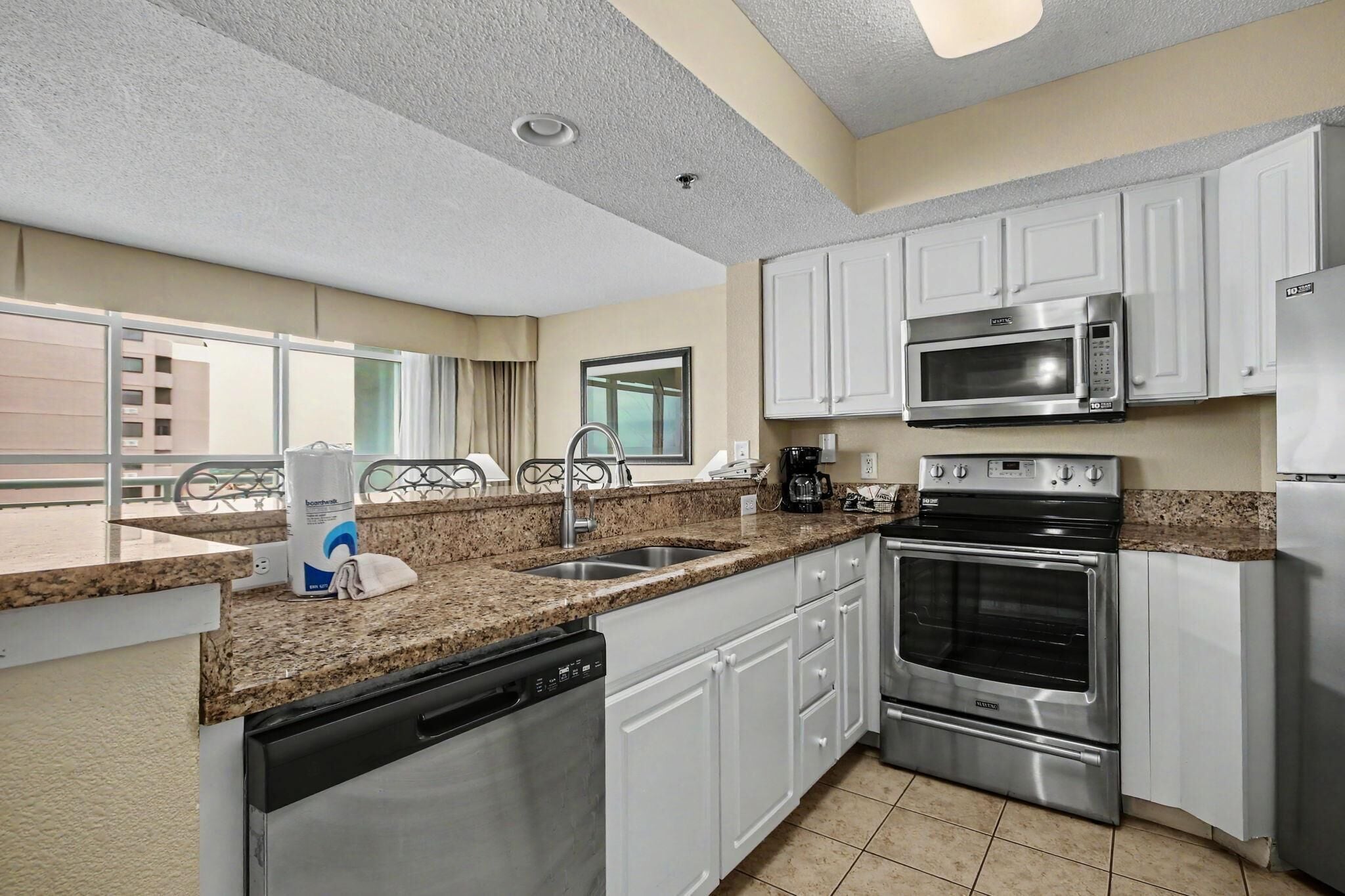 A modern kitchen with white cabinets, stainless steel appliances, a dishwasher, and a large window offering a view of the outside.