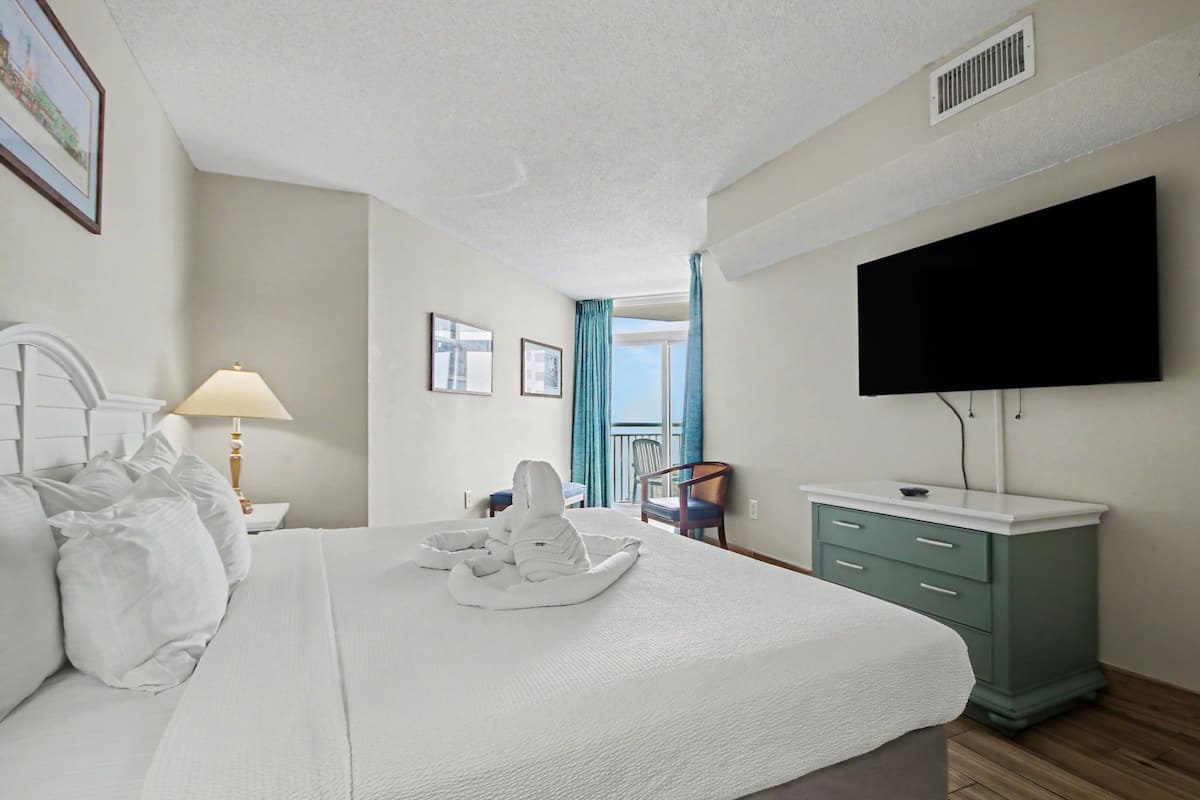 Habitación de hotel con cama, televisión, mesita de noche con lámpara, silla y vista al balcón.