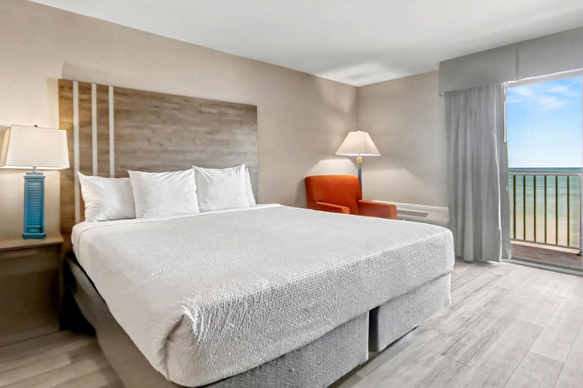 Habitación de hotel con una cama grande, dos mesitas de noche, una silla, un balcón con vistas y una ventana con cortinas.