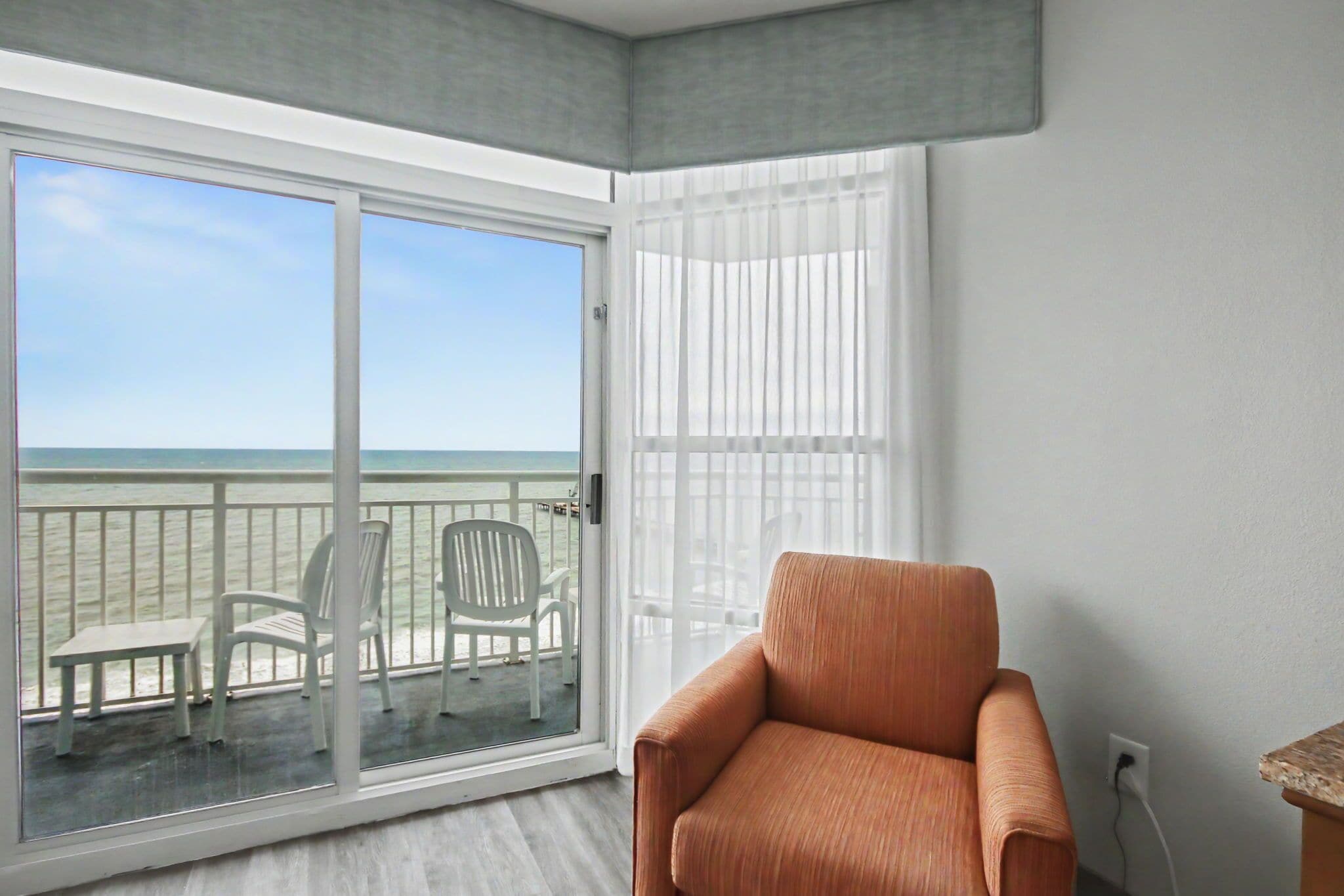 A room with a balcony, a white chair, a cushioned armchair, and a view of the ocean.