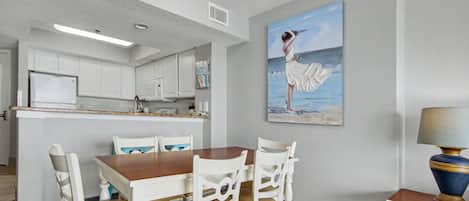 A dining area with a wooden table and white chairs, a painting of a woman on the beach, and a lamp on a side table.