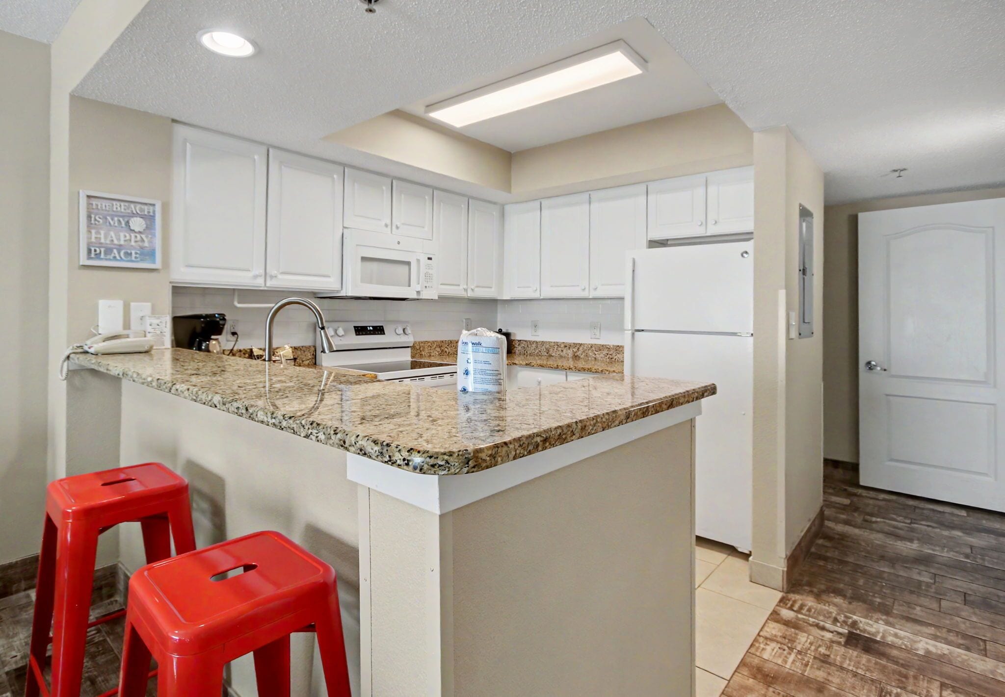 A kitchen with white cabinets, a granite countertop, a microwave, a refrigerator, and two red stools.