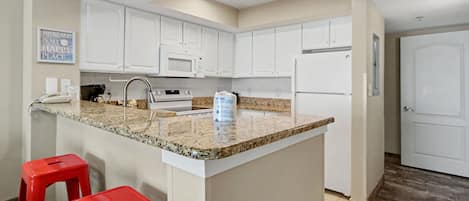 A kitchen with white cabinets, a granite countertop, a microwave, a refrigerator, and two red stools.