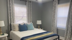 Un dormitorio con una cama, dos mesitas de noche, una lámpara en cada una y una ventana con persianas.