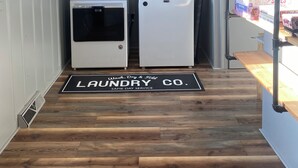 Un área de lavandería con dos lavadoras, piso de madera, una alfombra con el logo de 'Laundry Co.', una ventana con persianas y un estante con objetos.