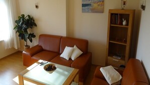 Uma sala de estar com um sofá marrom, uma mesa de centro de vidro, uma estante de livros e um vaso com planta.