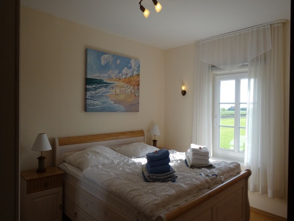 Ein Schlafzimmer mit einem Bett, einem Gemälde mit Strandansicht, einem Fenster mit Vorhängen und einem Nachttisch mit Lampe.