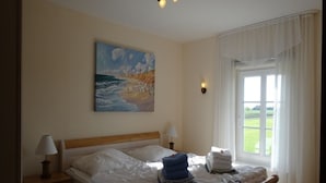 Quarto com cama, pintura de uma cena de praia, janela com cortinas e mesa de cabeceira com luminária.