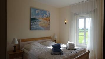 Ein Schlafzimmer mit einem Bett, einem Gemälde mit Strandansicht, einem Fenster mit Vorhängen und einem Nachttisch mit Lampe.