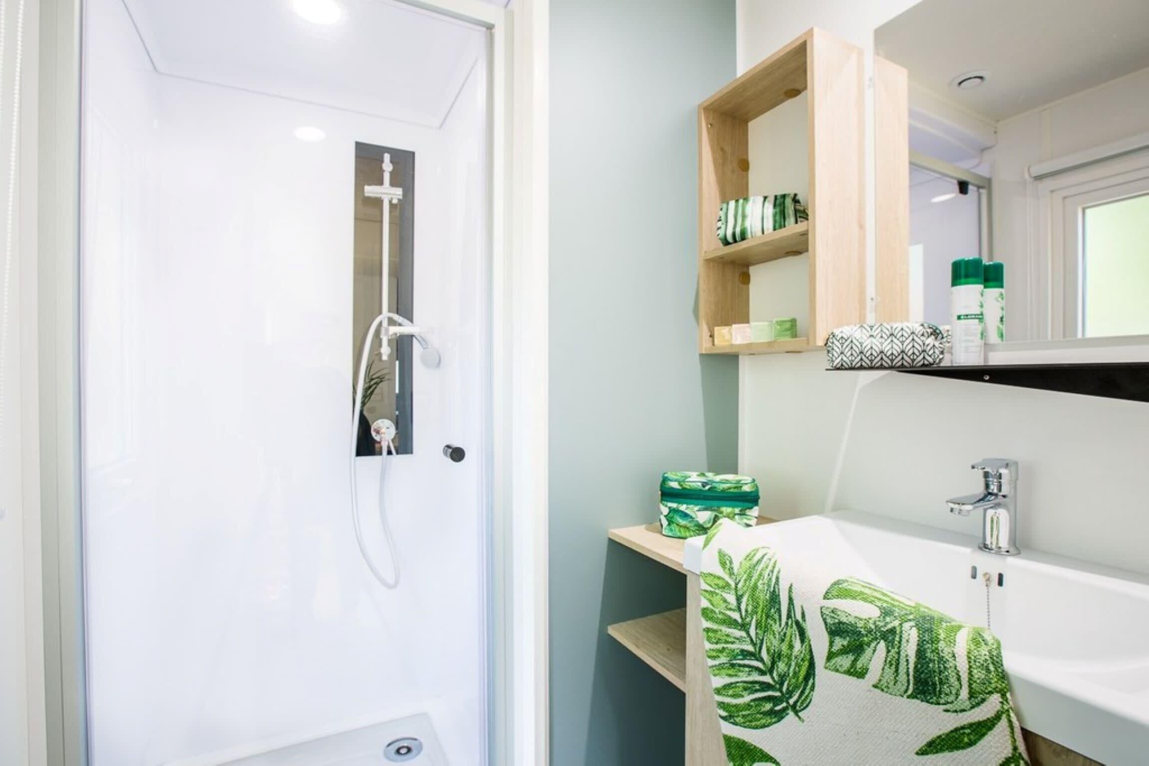 Un baño con puerta blanca, ducha, lavabo con espejo y estantes con artículos de aseo.