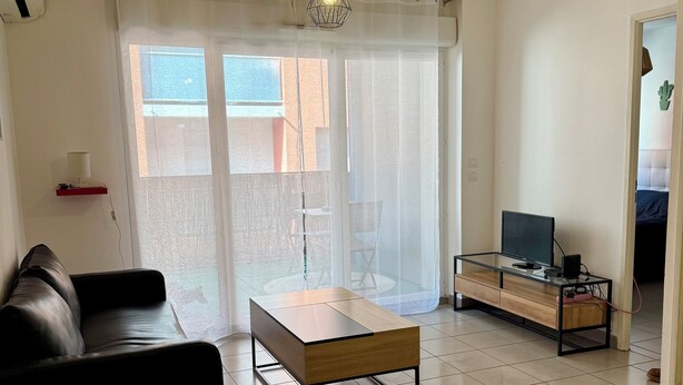 Een moderne woonkamer met een houten eettafel, een zwarte leren bank, een glazen salontafel en een televisie op een houten dressoir.