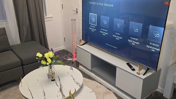 Una sala de estar moderna con un televisor de pantalla plana mostrando una interfaz de intensidad de señal de red, un sofá, una mesa de centro con un jarrón de flores y un reloj en la pared.