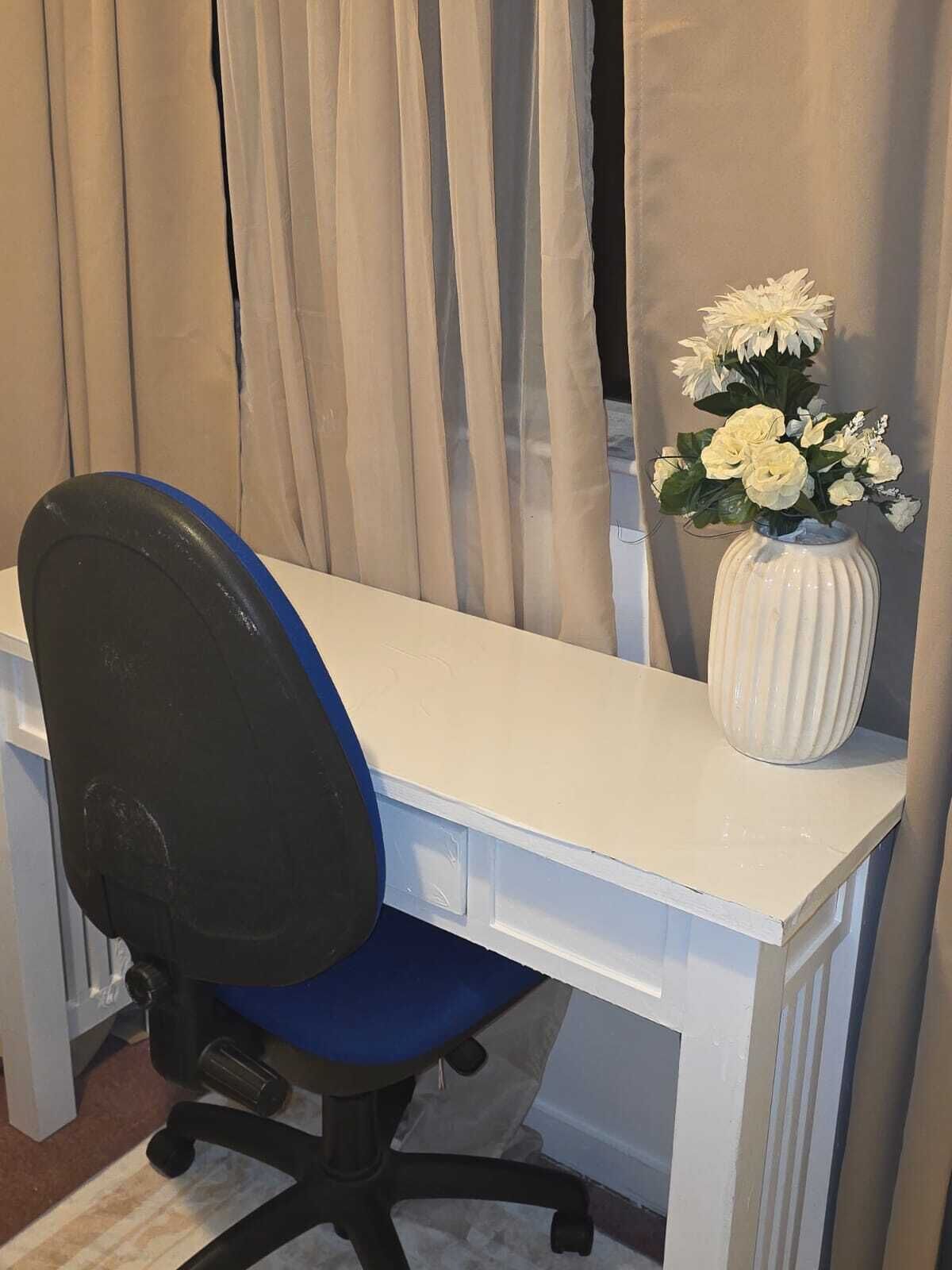 A white desk with a black office chair, a white vase with white flowers, and beige curtains.
