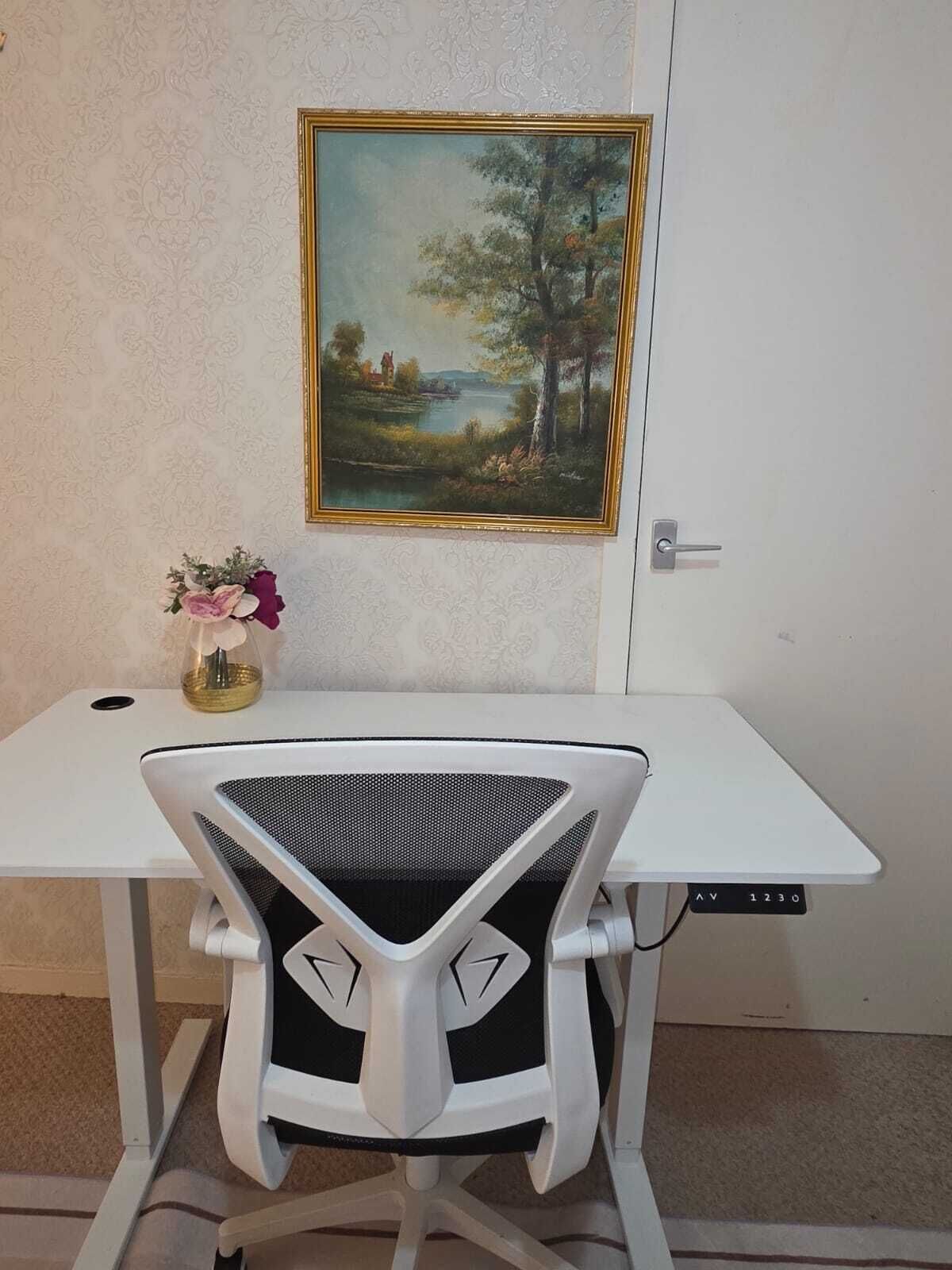 Ein weißer Schreibtisch, ein moderner Stuhl, ein gerahmtes Landschaftsbild und eine Vase mit Blumen an der Wand.