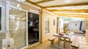 A wooden deck with a picnic table set up, a sliding glass door leading to a kitchen, and a small outdoor seating area. - CampingSpa - Confort 3 TV - 6p (33) (La Couarde-sur-Mer)