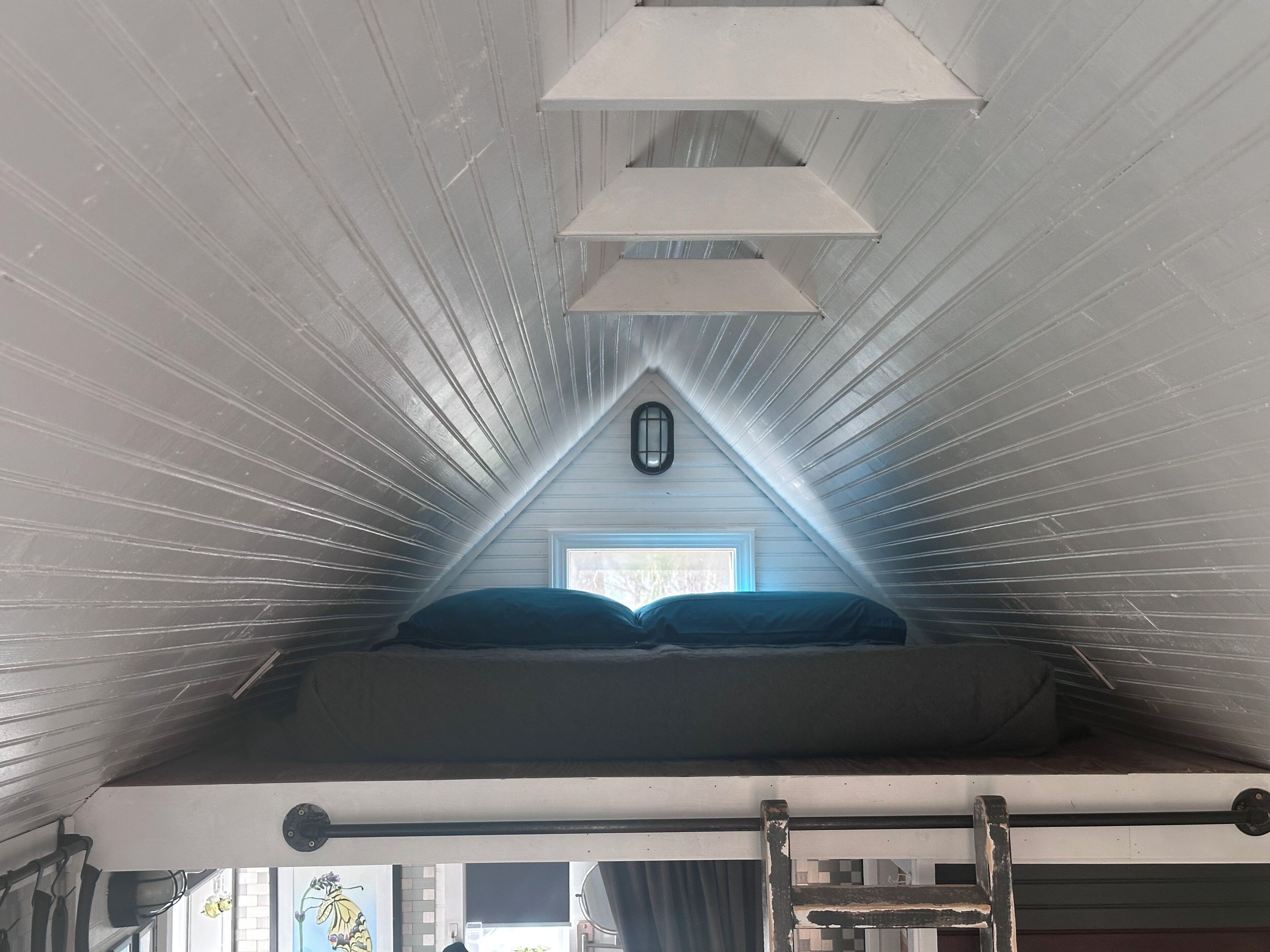 Ein Schlafzimmer im Loft-Stil mit schrägem Dach, einem Dachfenster, einem Bett mit Kissen und einer Himmelsleiter.