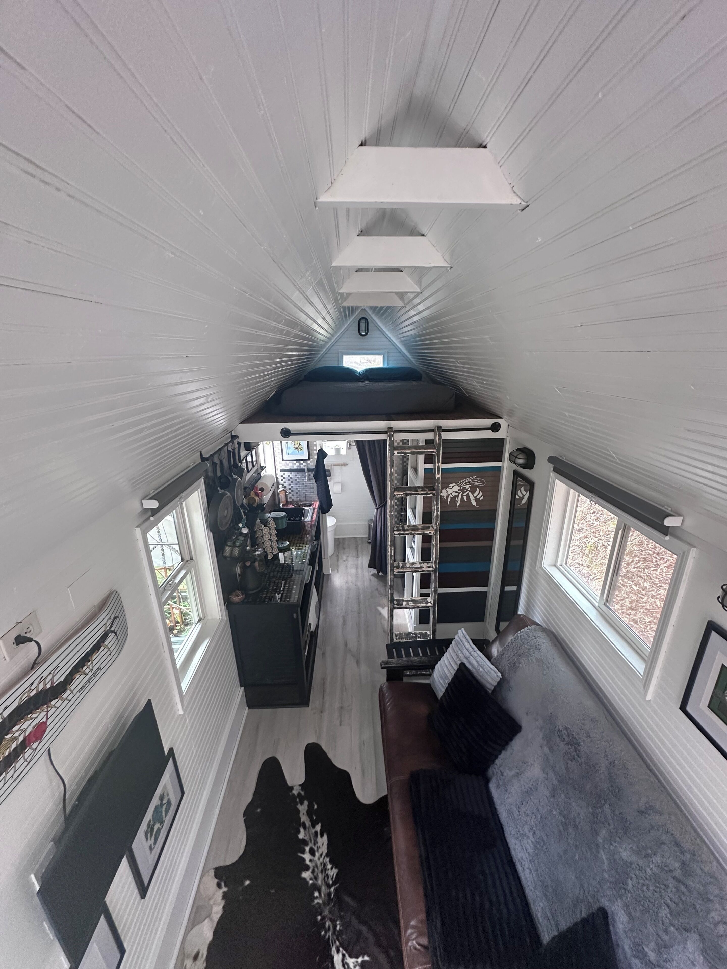 A compact, single-level interior with a loft bed, a seating area with a sofa and a chair, a kitchen area with a stove and counter, and a window providing natural light.