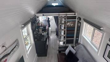 A compact, single-level interior with a loft bed, a seating area with a sofa and a chair, a kitchen area with a stove and counter, and a window providing natural light.