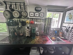 Ett kök med kakel på väggen bakom arbetsbänken, hängande kastruller, en kaffemaskin och ett fönster med utsikt över grönskan.