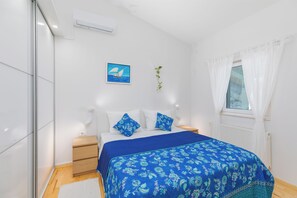 Un dormitorio con cama, mesitas de noche, una ventana con cortinas, un aire acondicionado de pared y un cuadro enmarcado.