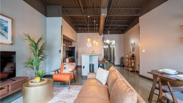 A modern living room with a sofa, armchairs, a coffee table, and a dining table. There is a kitchen in the background with a refrigerator and a sink. - Sleek Industrial Loft in Short North Near OSU with King Beds & Fast WiFi (Columbus)