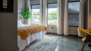 A bedroom with a bed, a chair, a plant, and large windows with curtains. - Sleek Industrial Loft in Short North Near OSU with King Beds & Fast WiFi (Columbus)