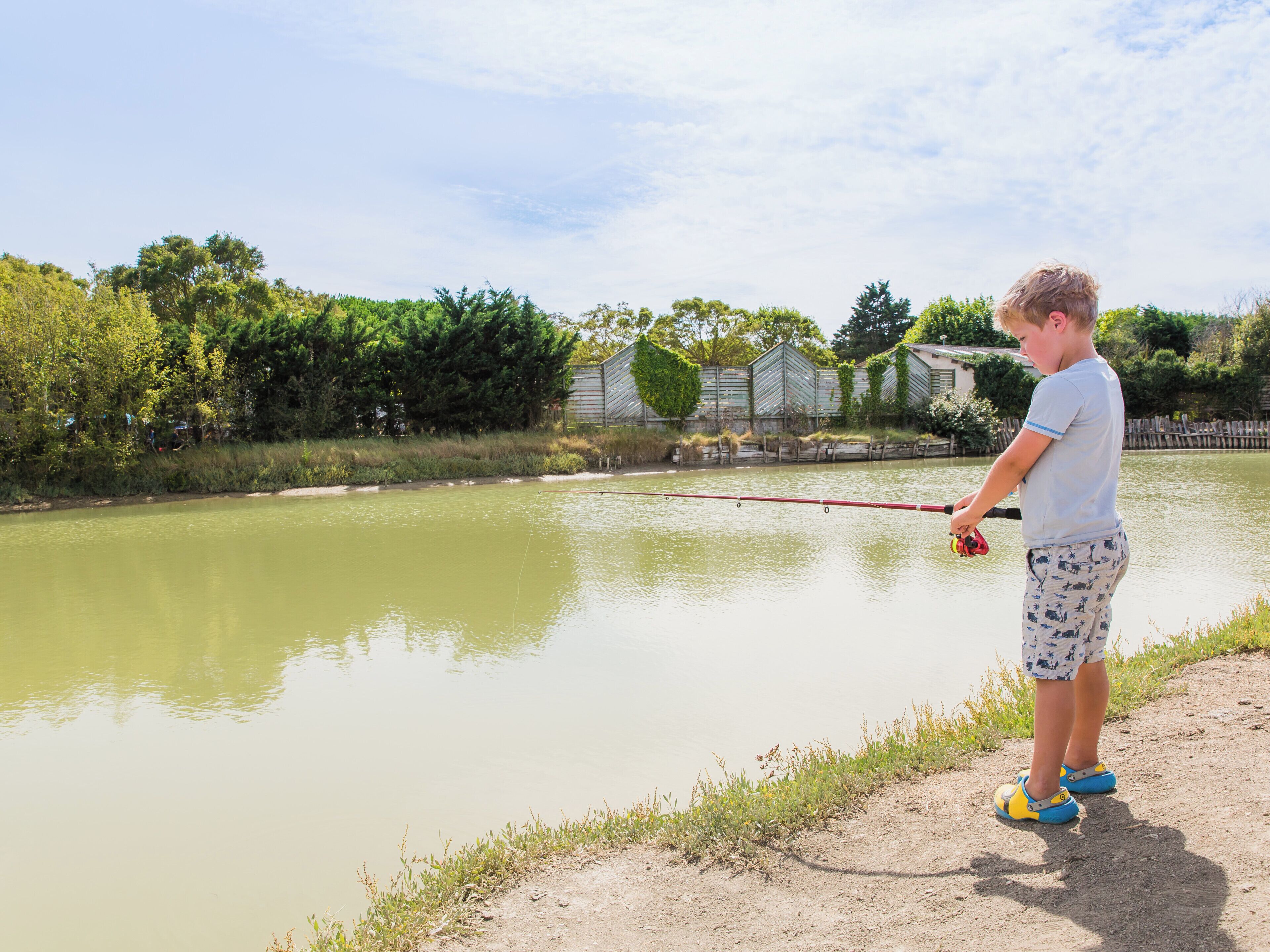 Un enfant est en train de pêcher au bord d’un étang paisible, avec une clôture en bois et de la verdure en arrière-plan.