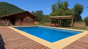 Una terraza de madera con piscina rectangular, una cabaña de madera y un área de estar cubierta con dos sillones reclinables.