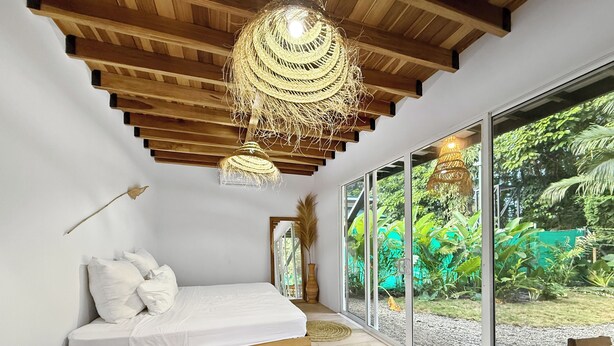 Ein Schlafzimmer mit einem hölzernen Bett, einer gewebten Deckenleuchte und einem großen Fenster mit Blick auf Grünflächen.