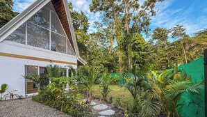 Ein modernes Haus mit einem dreieckigen Dach, großen Fenstern und einem Garten mit Kiesweg und tropischen Pflanzen.