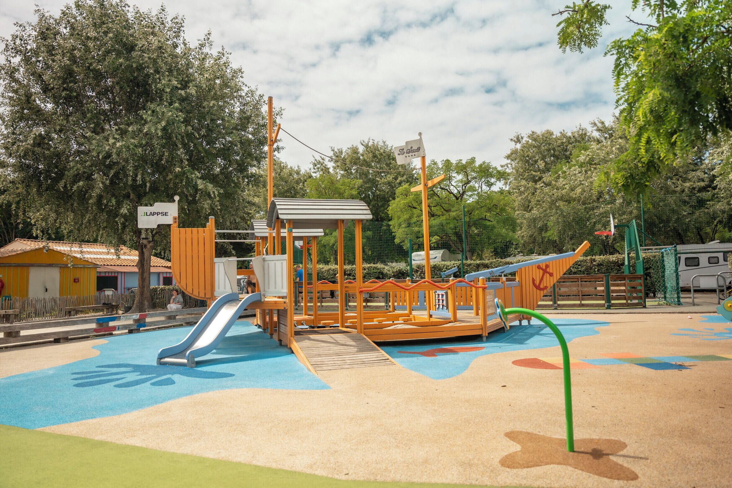 A playground with slides and climbing structures, surrounded by trees and a fenced area.