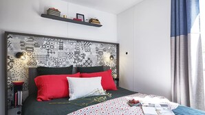Ett modernt sovrum med en mönstrad sänggavel, röda och vita kuddar, ett blått överkast och ett nattduksbord med böcker och en lampa.