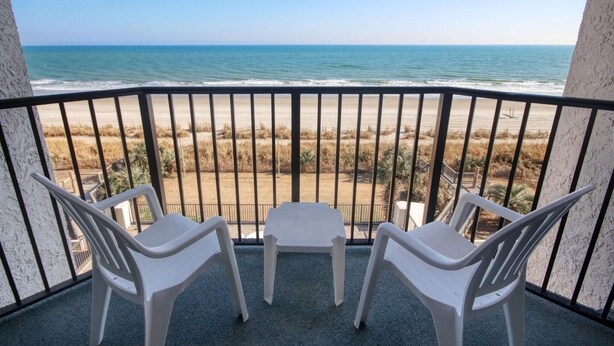 Un balcon avec deux chaises en plastique blanc et une petite table, d’où l’on a une vue dégagée sur la plage et l’océan.