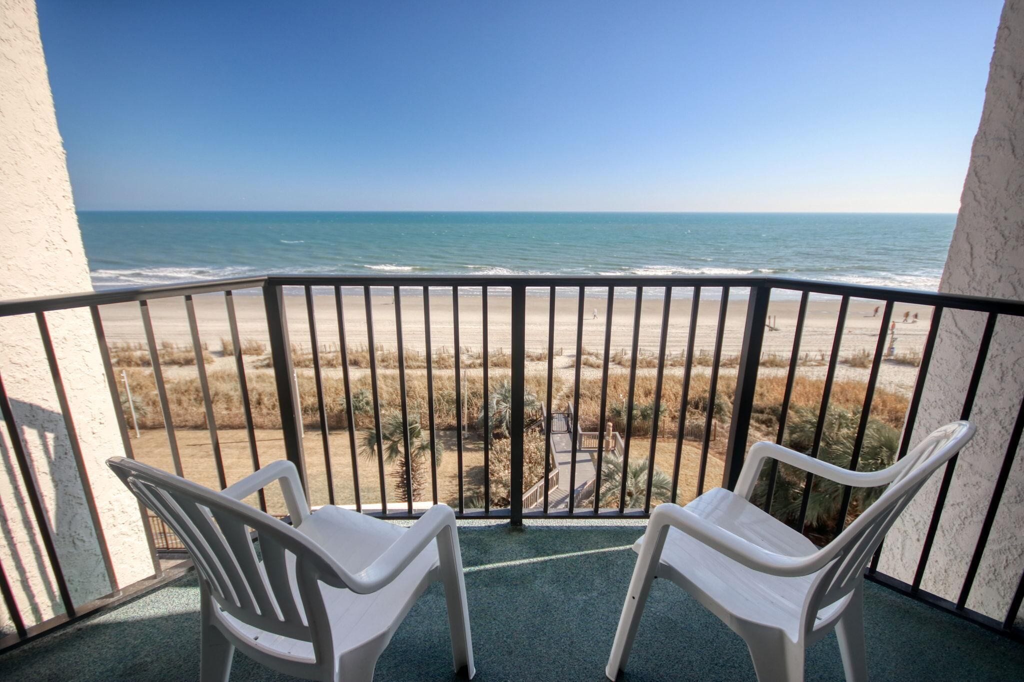 Un balcon avec deux chaises en plastique blanches donnant sur une plage, sous un ciel dégagé.