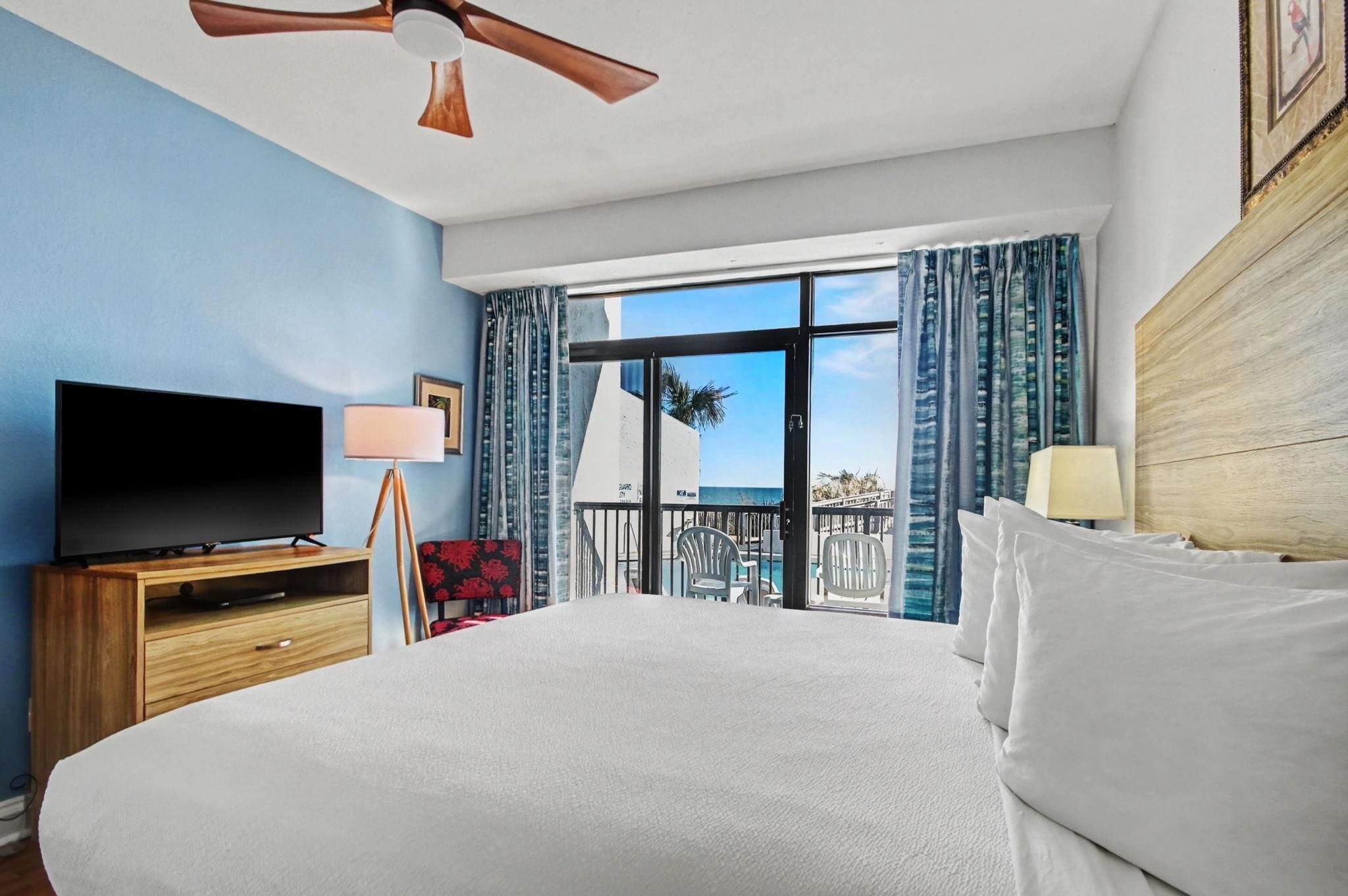A hotel room with a bed, a TV, a ceiling fan, a balcony with a view of the ocean, and a window with curtains.