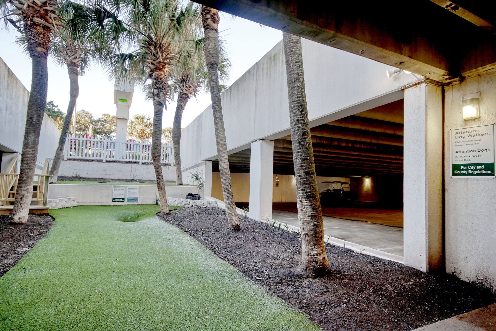 A courtyard with artificial grass, palm trees, and a covered area with a sign about dog walking regulations.