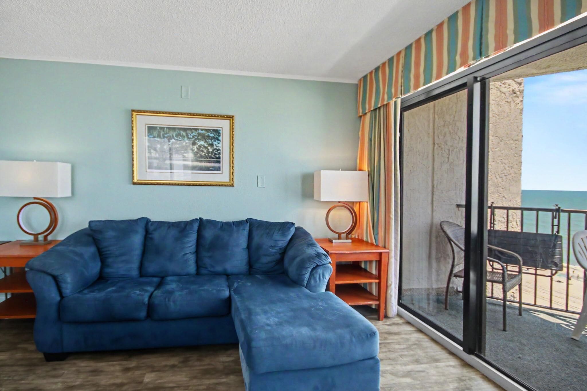 A room with a blue sectional sofa, two side tables with lamps, a framed picture on the wall, and a view of the sea through a sliding glass door.