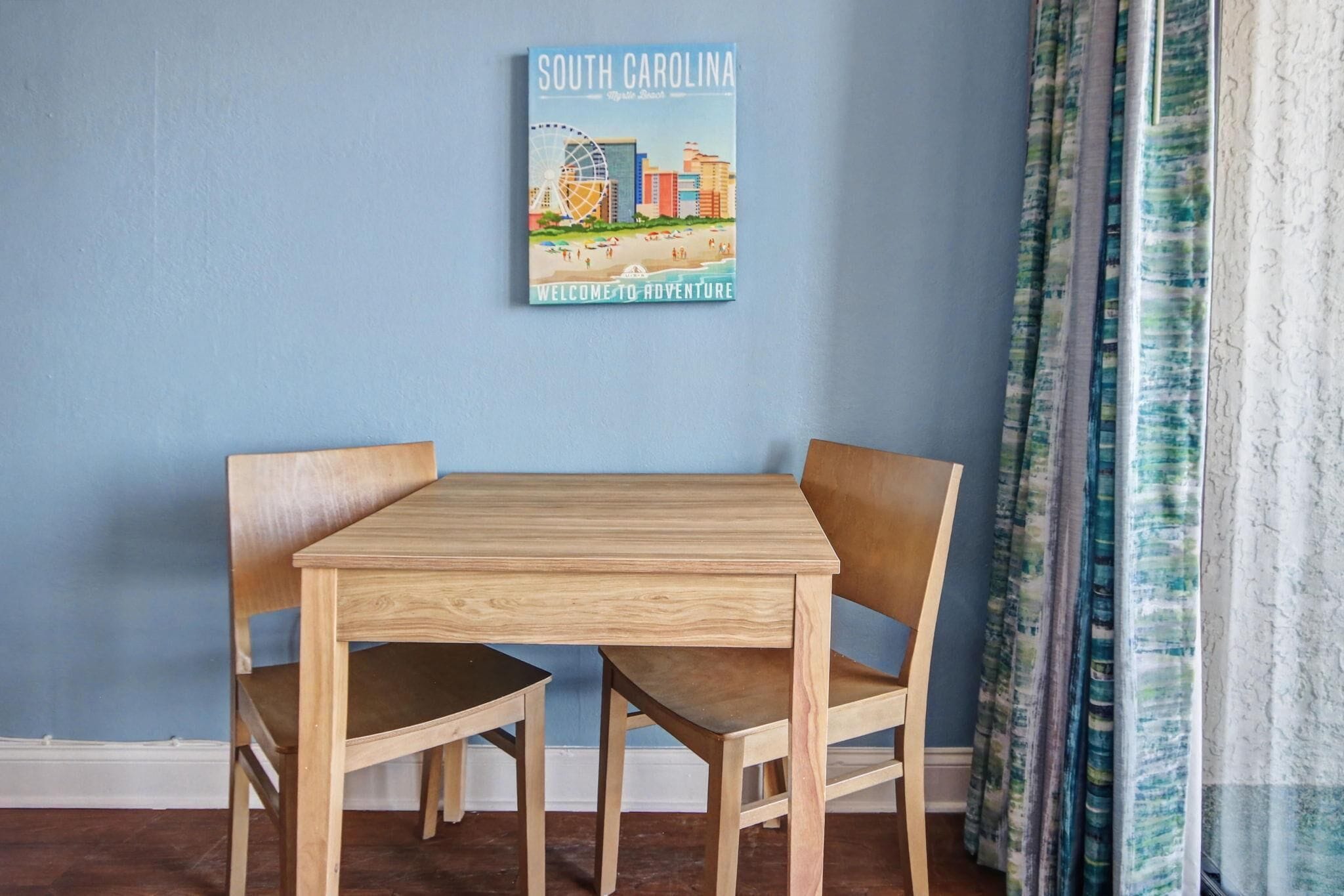 Un coin repas avec une table et des chaises en bois, une affiche représentant un paysage urbain accrochée au mur et une fenêtre avec des rideaux.