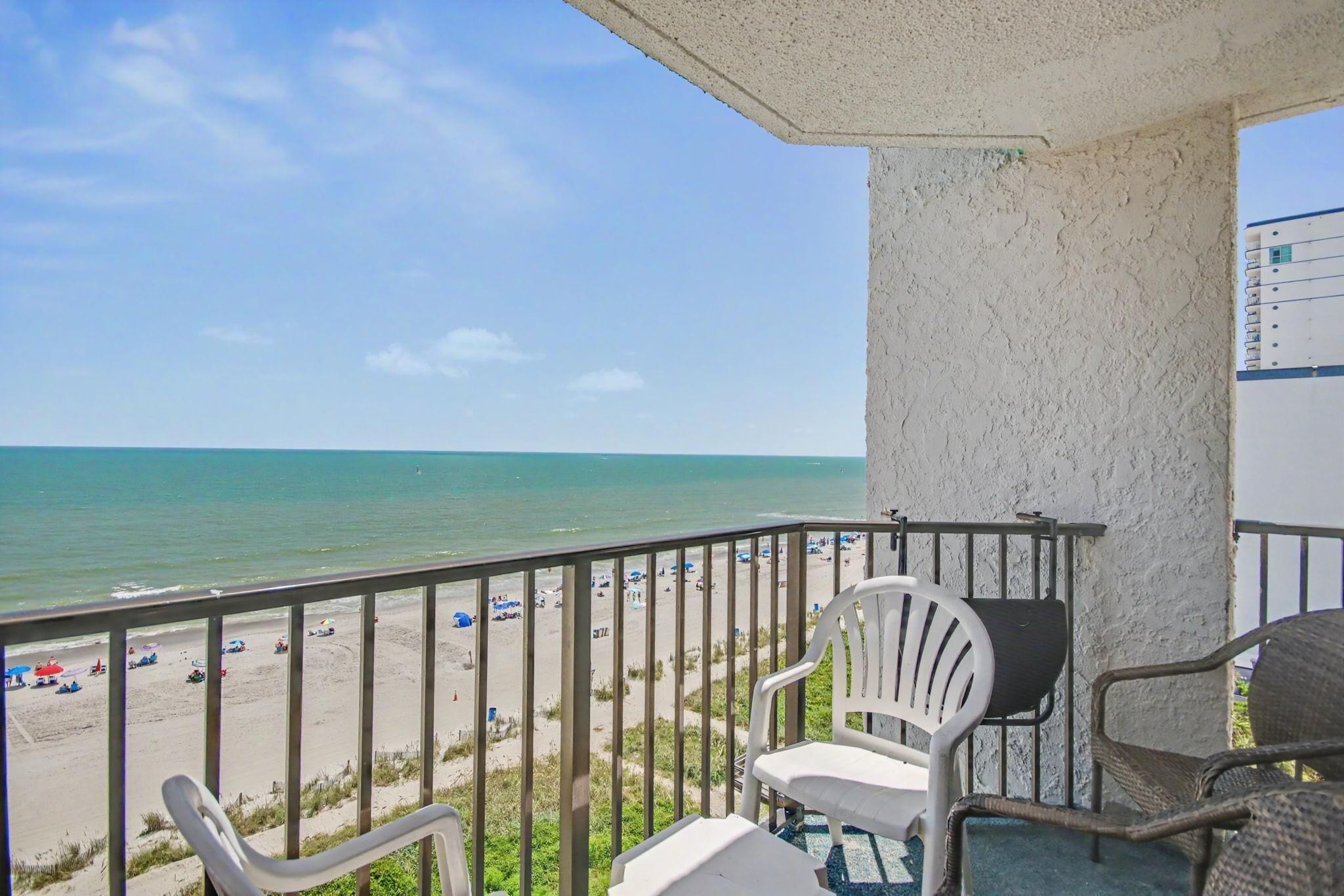 A balcony with a view of a beach and ocean. There are two white chairs and a table.