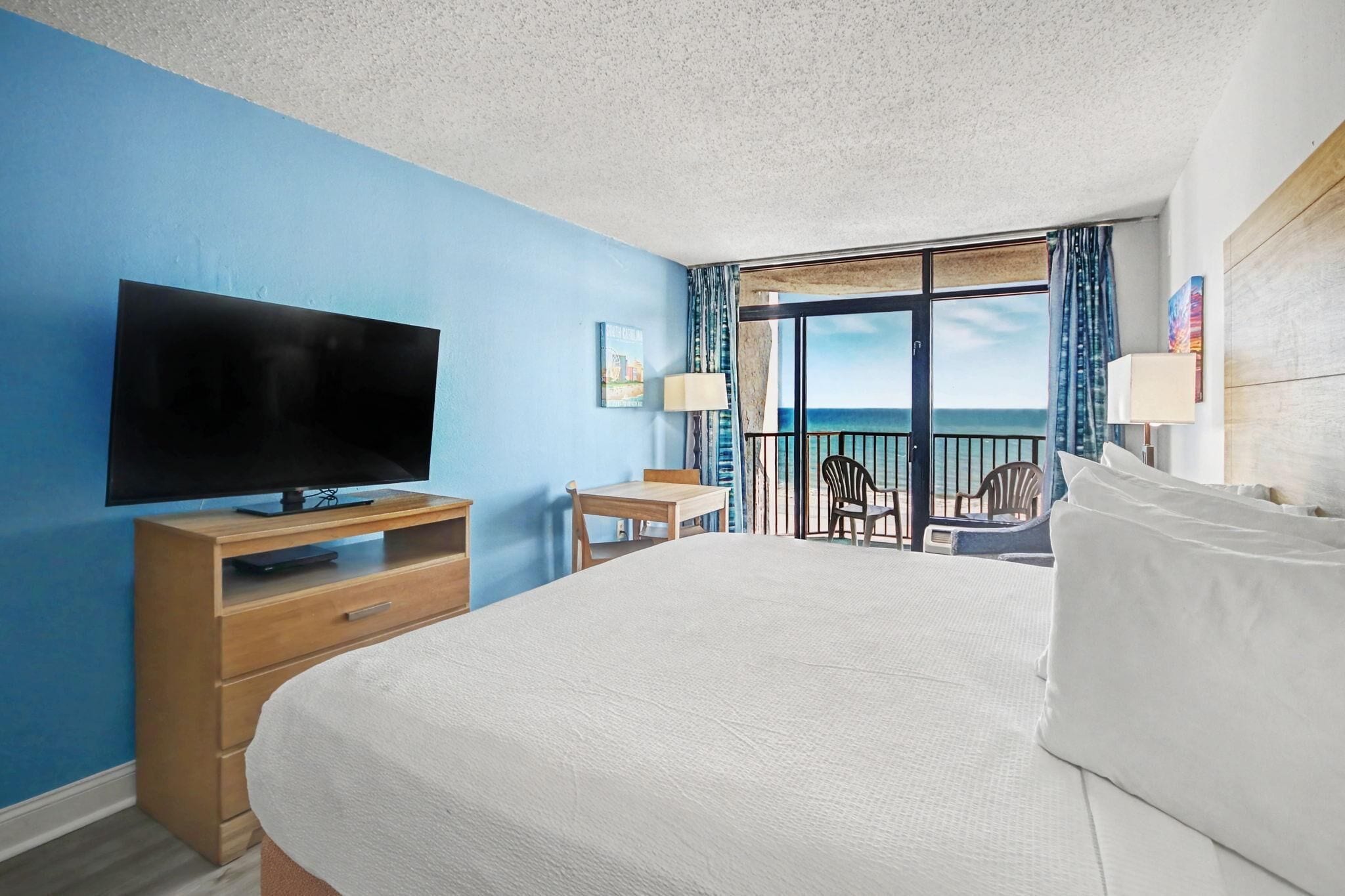 A hotel room with a large bed, a TV, a desk, and a balcony with a view of the ocean.
