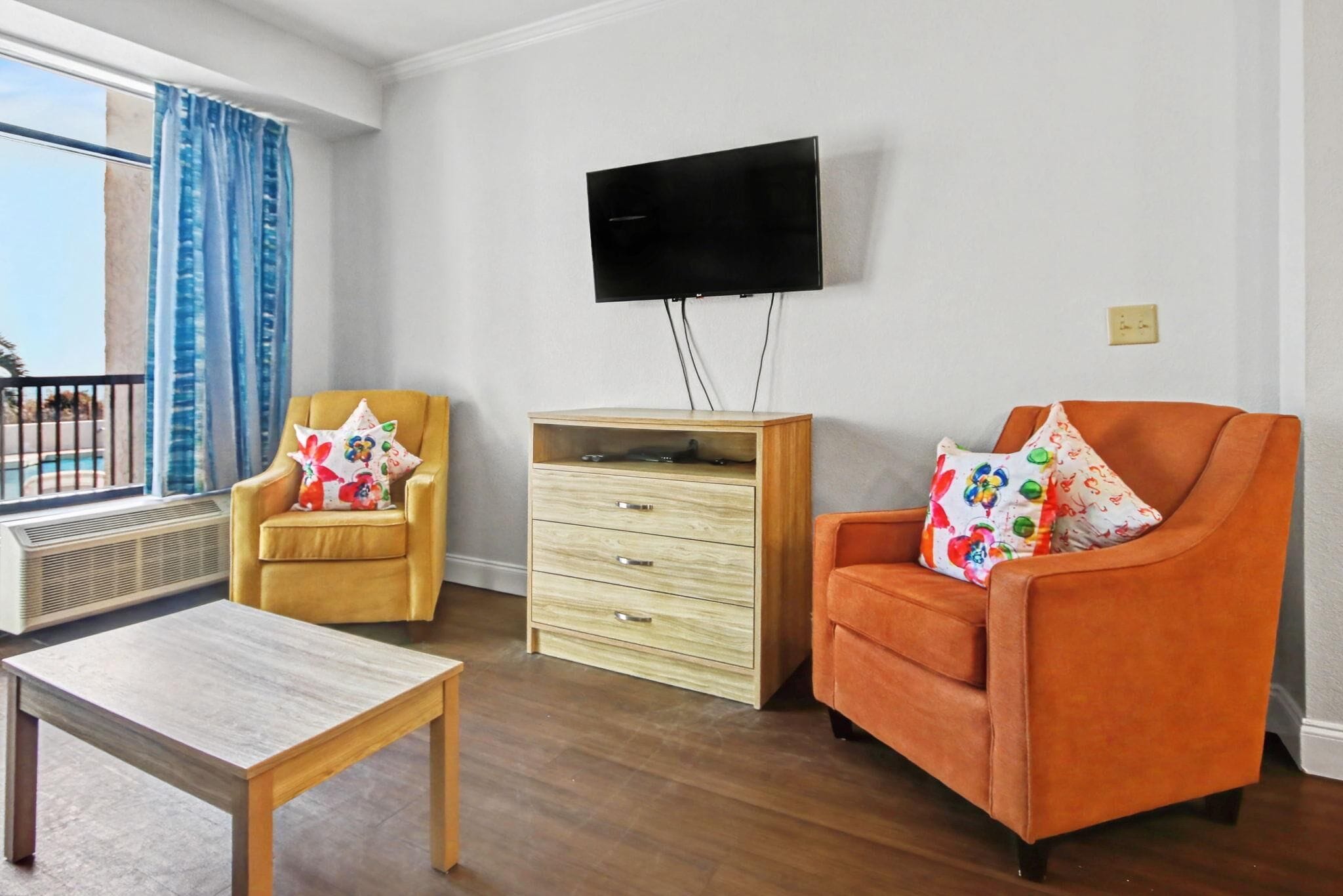Een kamer met een houten salontafel, twee fauteuils, een aan de muur bevestigde televisie en een raam met blauwe gordijnen.