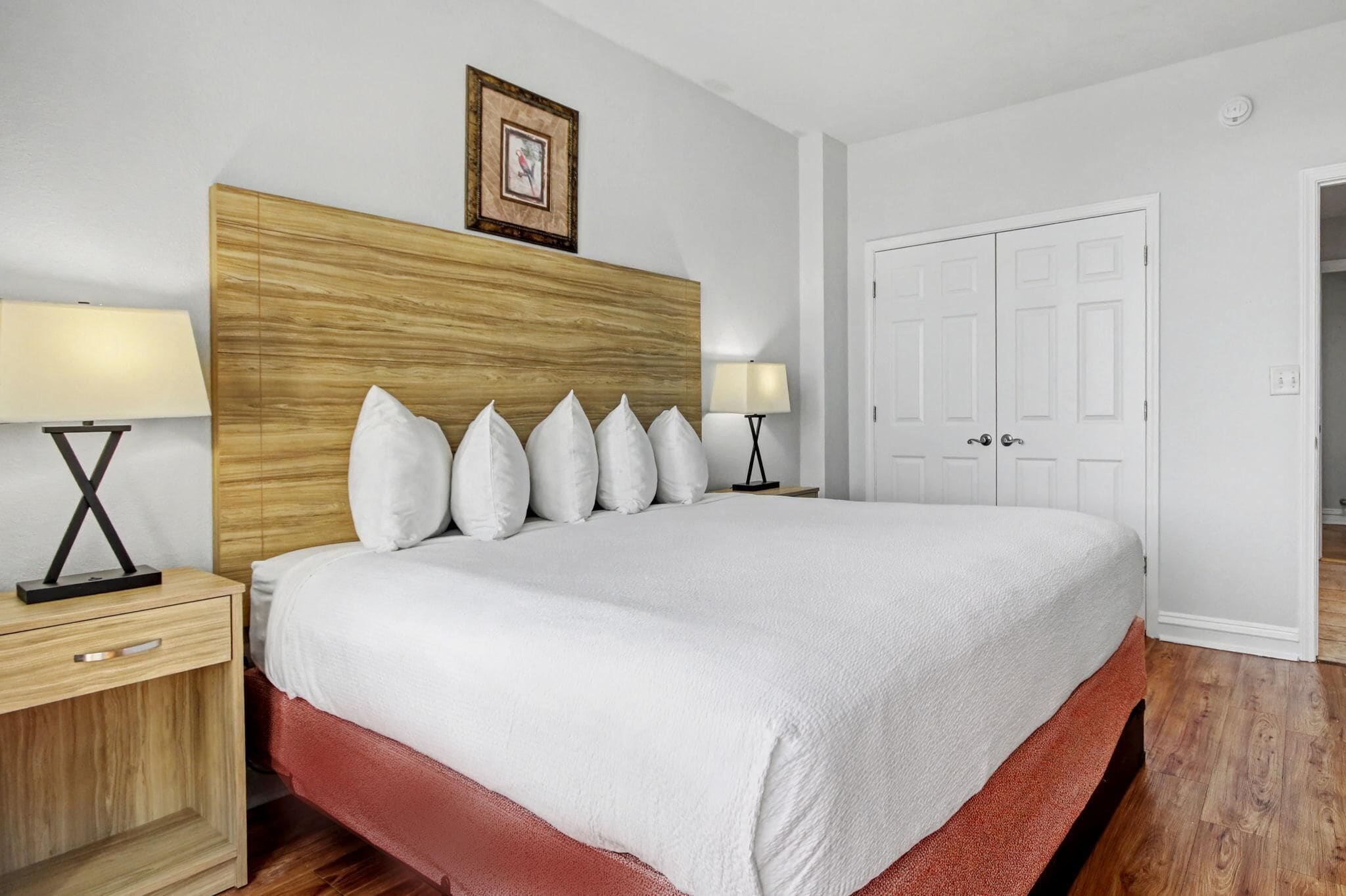 Habitación de hotel con cabecera de madera, cama grande con ropa blanca, dos mesitas de noche con lámparas y una puerta blanca.