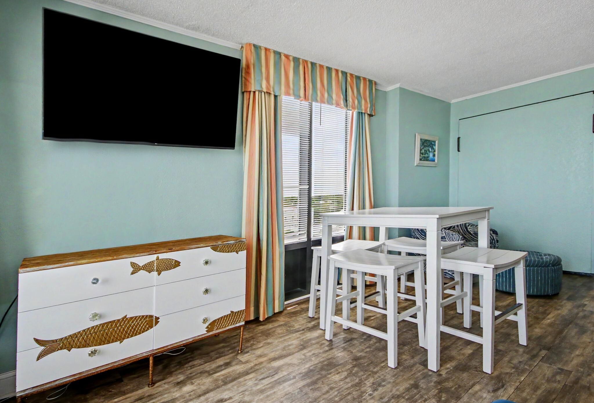 Ein Zimmer mit weißem Schminktisch, einem an der Wand befestigten Fernseher, einem Fenster mit gestreiften Vorhängen und einem kleinen Tisch mit zwei Stühlen.