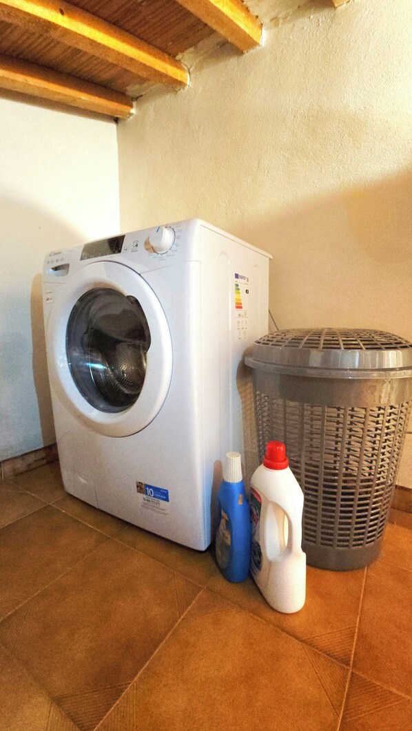 A front-loading washing machine, a laundry basket, and two bottles of detergent on a tiled floor.