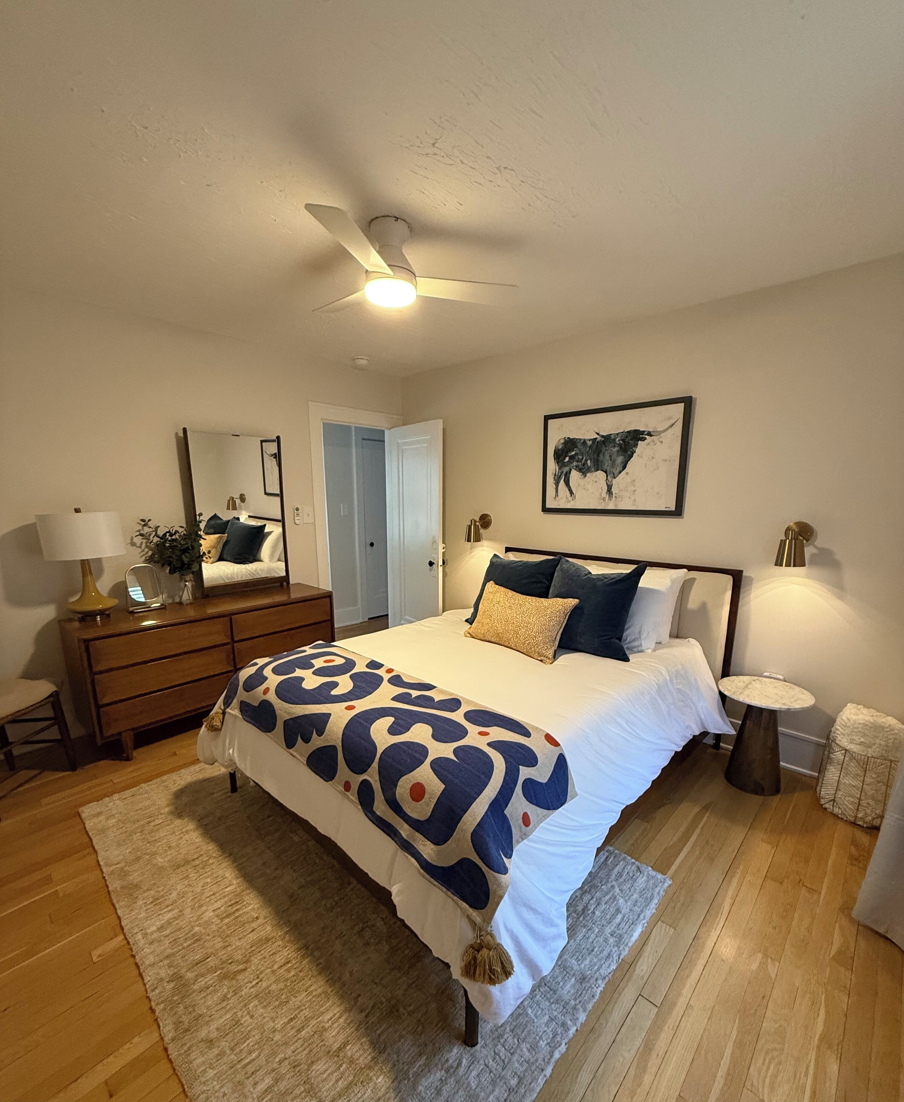 A bedroom with a bed, bedside tables, a dresser, a ceiling fan, a mirror, and a wall-mounted artwork.