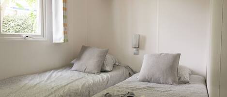 A bedroom with two single beds, a window with a view of greenery, a wall-mounted shelf, and a wall light.