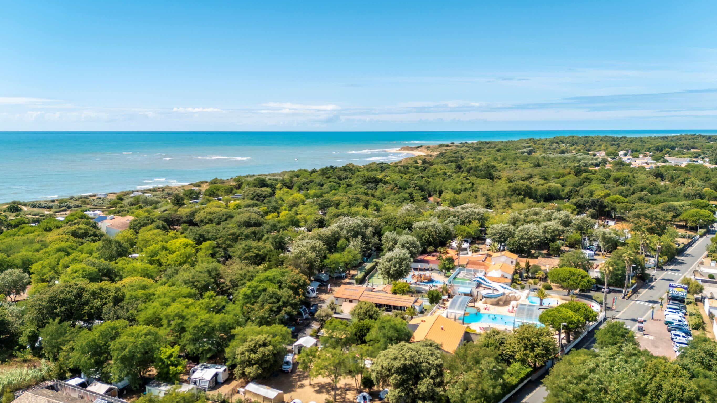 An aerial view of a coastal area with dense greenery, a swimming pool complex, and a beach in the background.