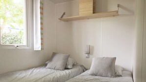 Un dormitorio con dos camas individuales, una ventana con vistas a zonas verdes, un estante montado en la pared y una lámpara de pared.