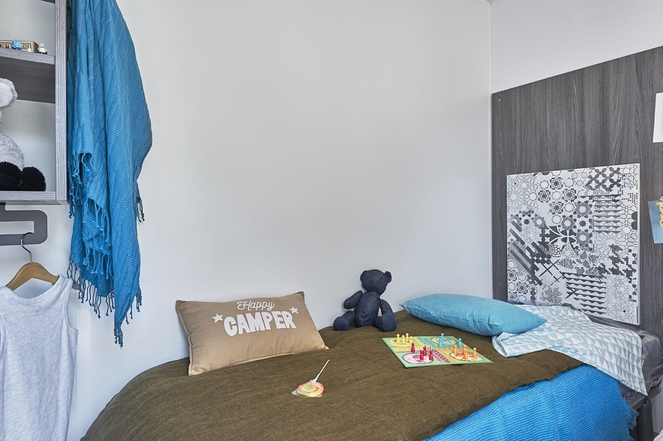 A bedroom with a bed, a teddy bear, a pillow with "Happy Camper" text, a lollipop, and a wall art piece.